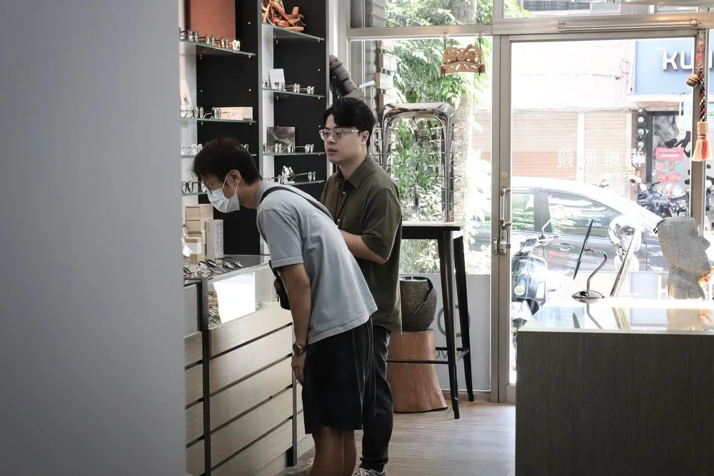 兩位顧客彎腰檢視玻璃展示櫃內的鏡框,門外是騎樓街景