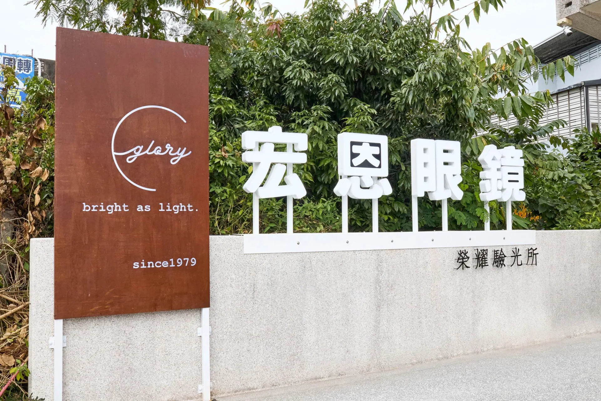 店前招牌：鏽鐵板上的 glory 標誌與 since1979，右側白色立體字「宏恩眼鏡」，牆面標示榮耀驗光所