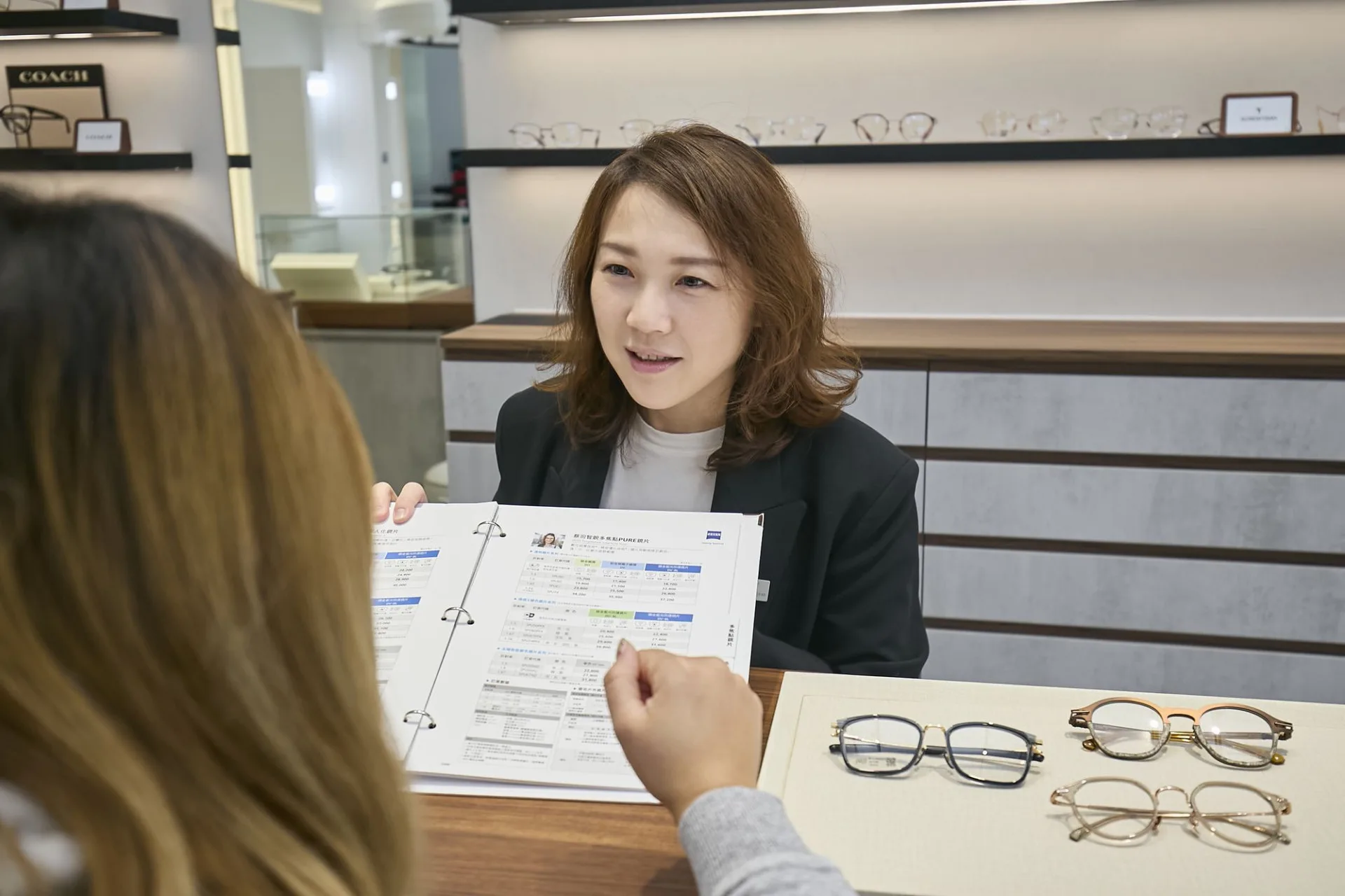 門市人員向顧客翻閱蔡司鏡片說明資料，桌面並排三副鏡框