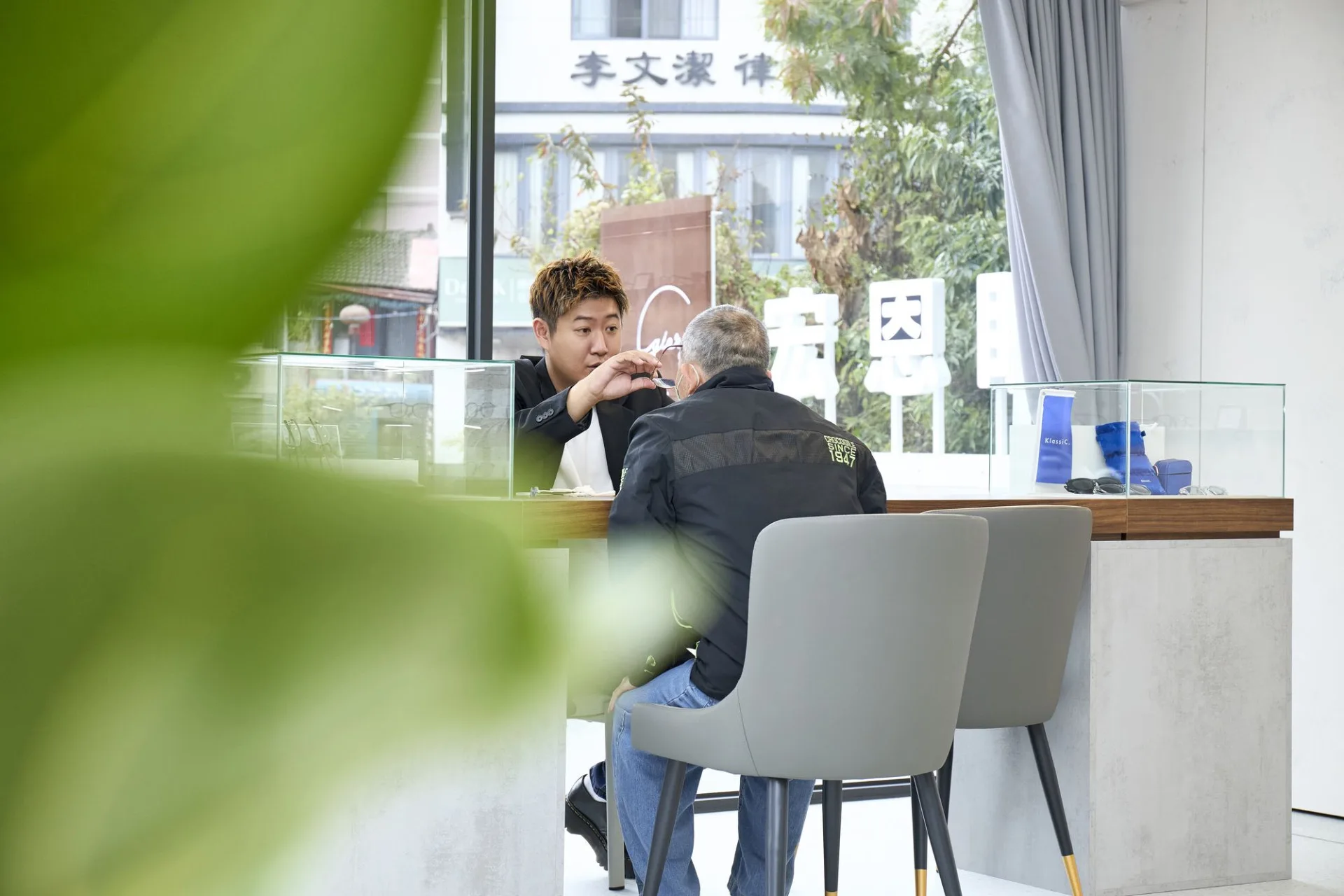 門市人員在諮詢桌前為長者顧客調整眼鏡，窗外可見宏恩眼鏡立體招牌
