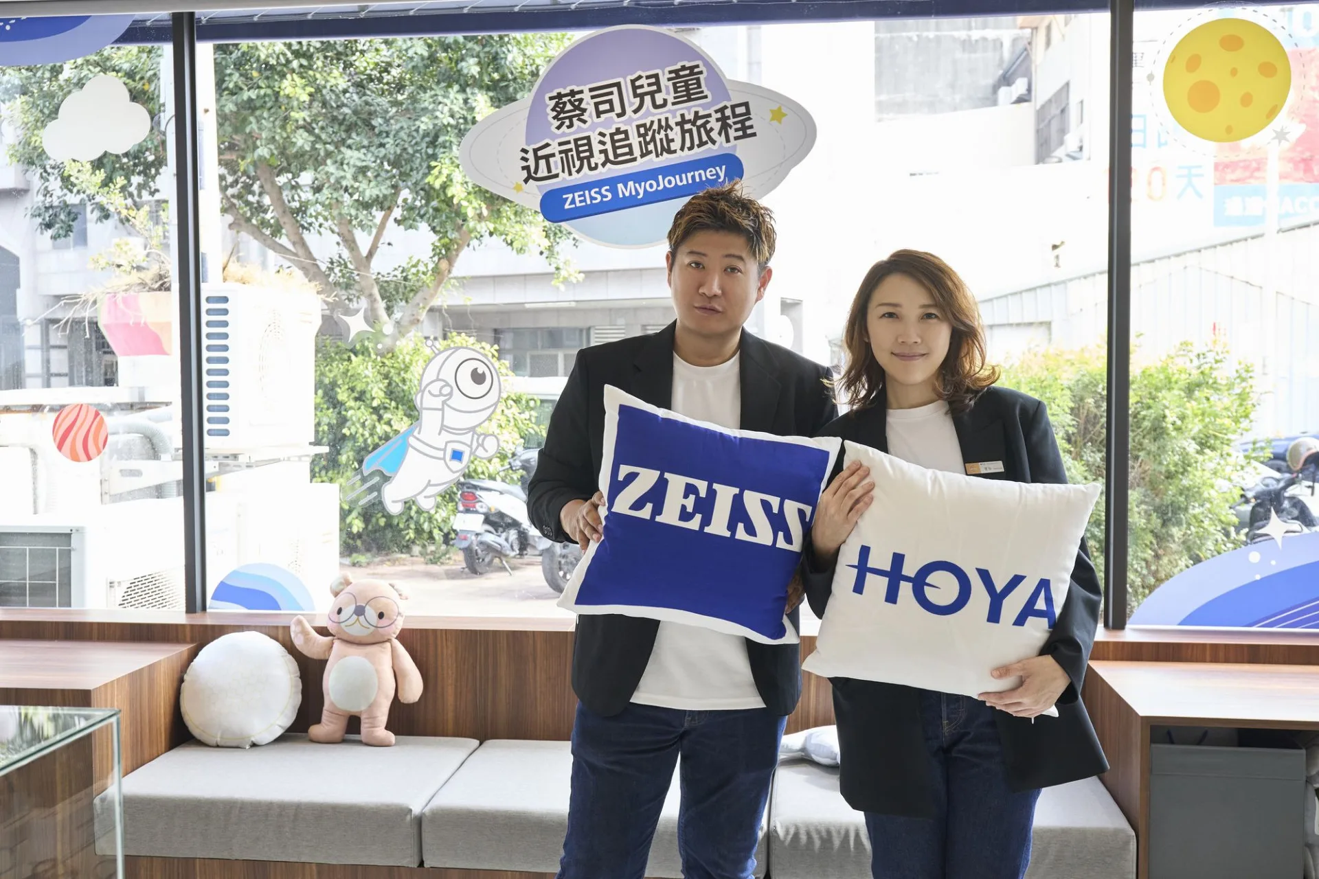 兩位主理人手持 ZEISS 與 HOYA 抱枕，站在蔡司兒童近視追蹤旅程窗貼前合影