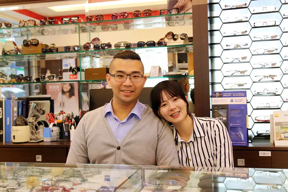 葉育仁夫妻在玻璃櫃檯後合影,身後是墨鏡陳列櫃與蜂巢格金屬框牆