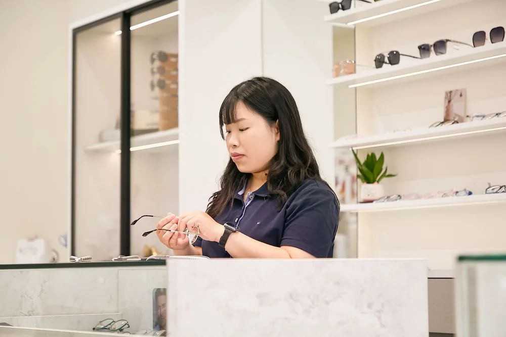 Stella 在櫃檯前低頭檢視手中的金屬細框眼鏡