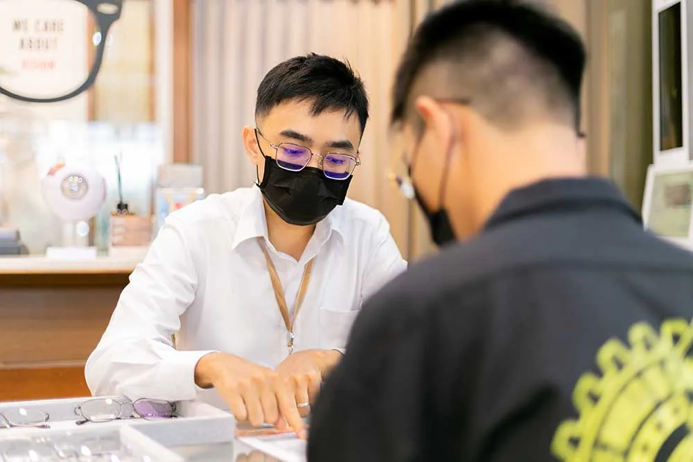 戴眼鏡的店員俯身指著玻璃櫃檯上的鏡框向顧客說明