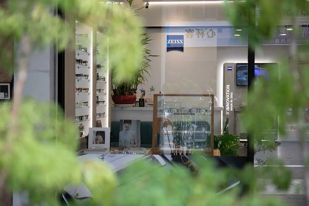 從門外綠葉間望向店內：ZEISS 燈箱、鏡框陳列牆與櫃檯形象照