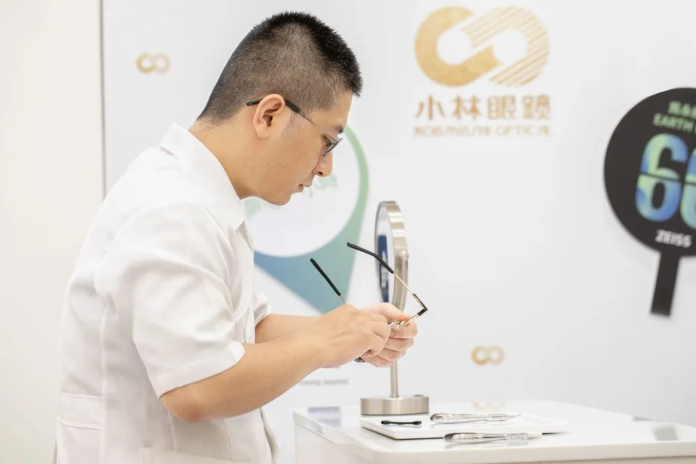 白袍配鏡師於小林眼鏡標誌牆前低頭調整金屬鏡框